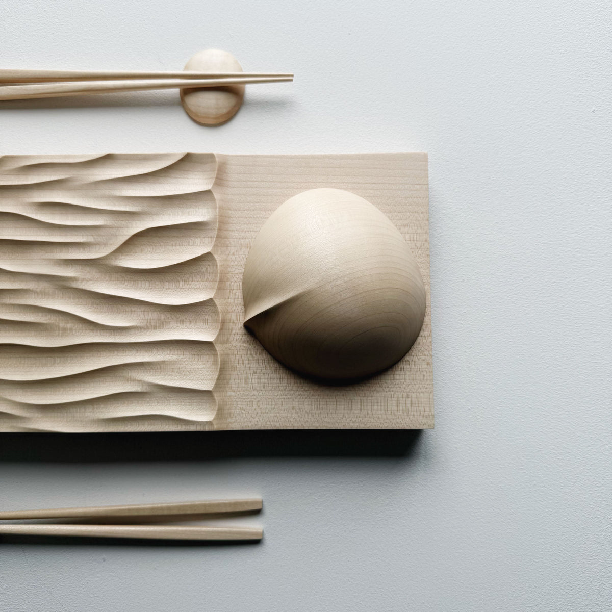 Making Waves sycamore board, with oval bowl, chopsticks and pebble rests in sycamore on a light background.