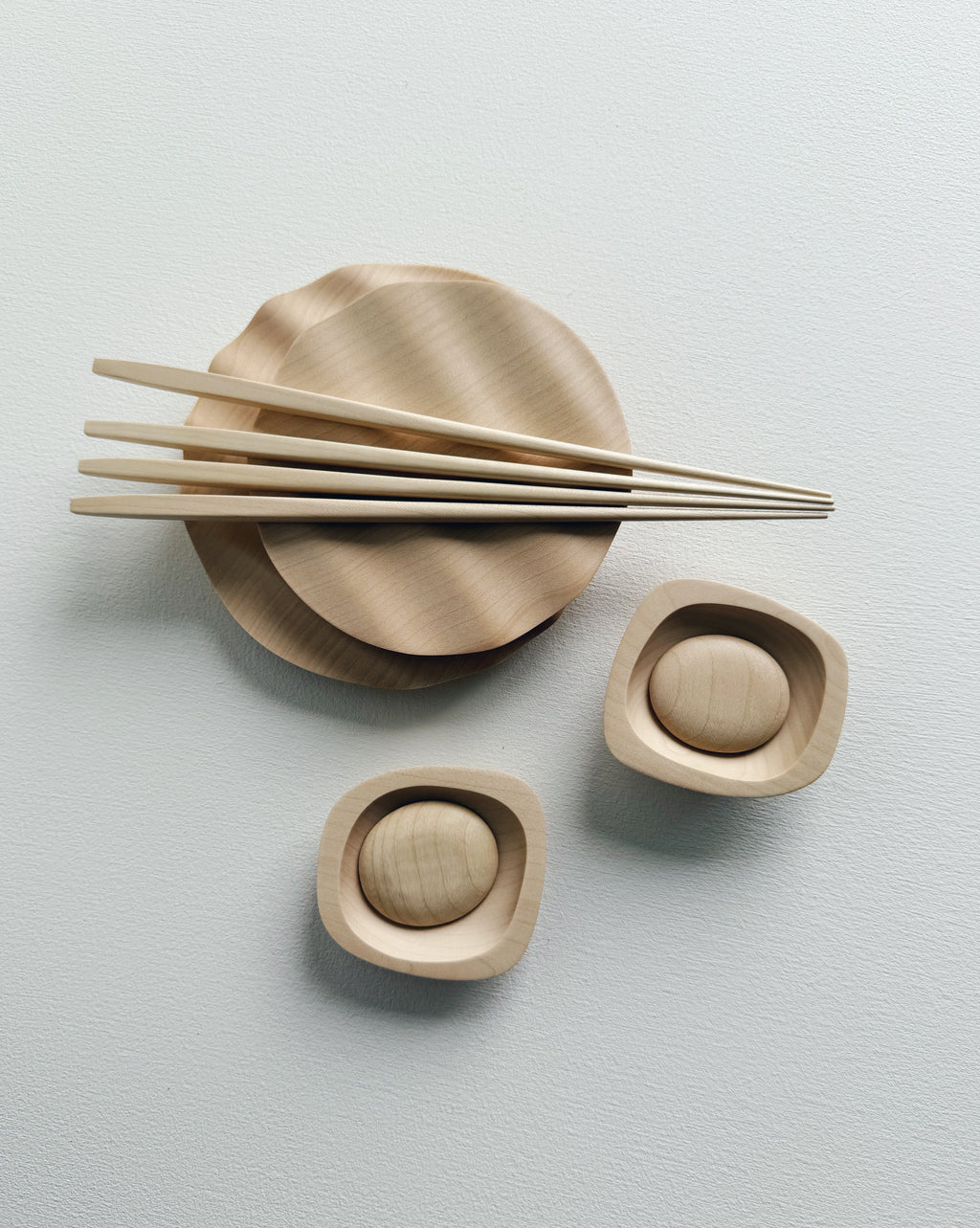 Wavy plates, plinth bowls, chopsticks and pebble rests in sycamore on a light background.