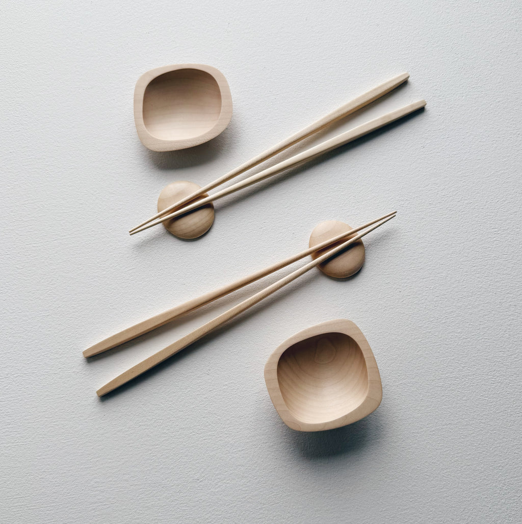Wavy plates, plinth bowls, chopsticks and pebble rests in sycamore on a light background.