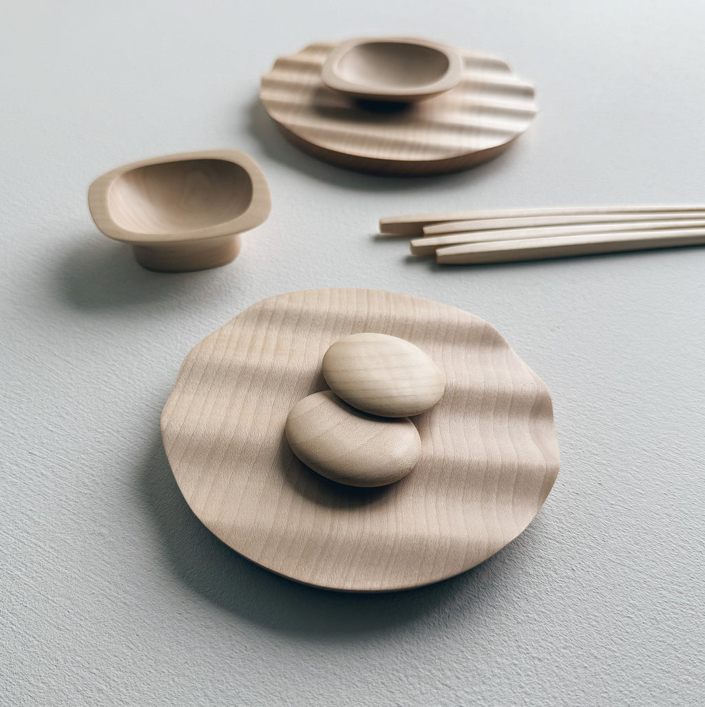Wavy plates, plinth bowls, chopsticks and pebble rests in sycamore on a light background.