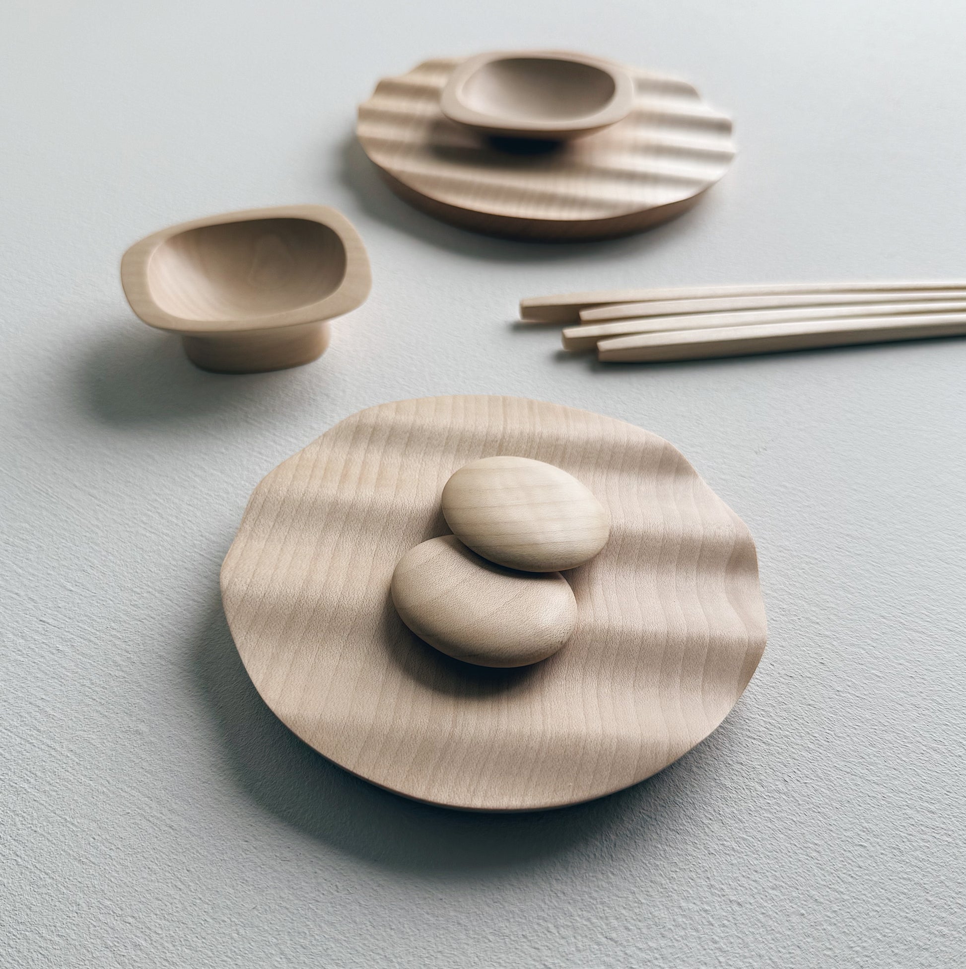 Wavy plates, plinth bowls, chopsticks and pebble rests in sycamore on a light background.