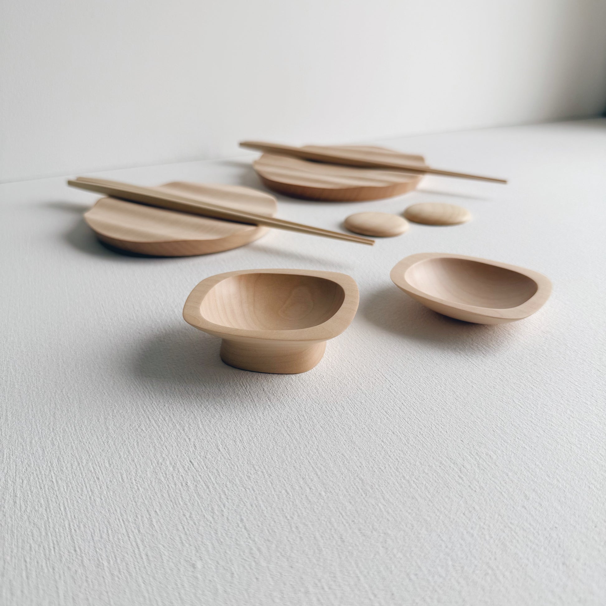 Wavy plates, plinth bowls, chopsticks and pebble rests in sycamore on a light background.