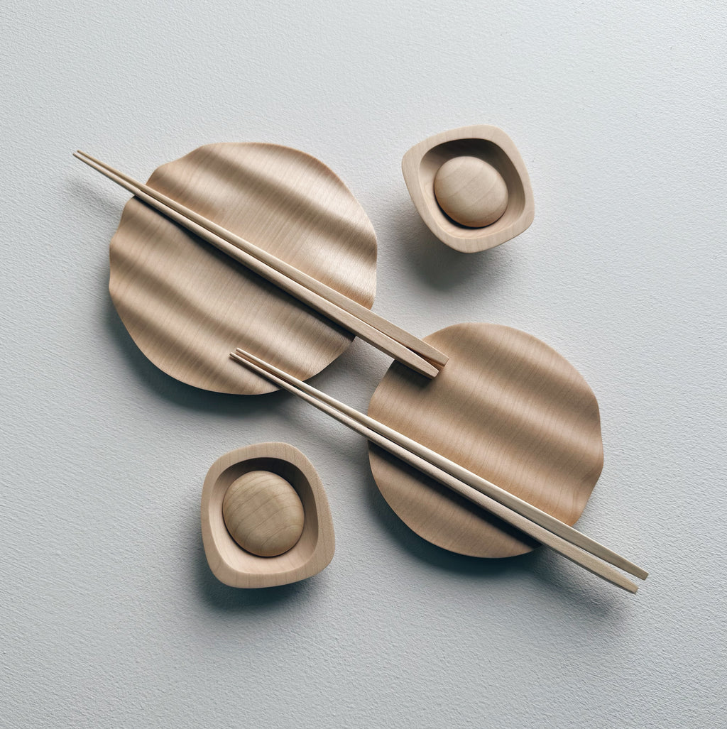 Wavy plates, plinth bowls, chopsticks and pebble rests in sycamore on a light background.